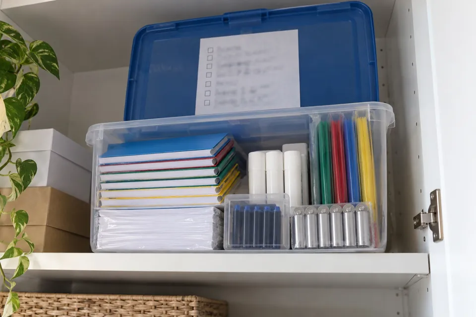 Bac de rangement transparent à couvercle bleu sur la tablette haute d’un placard, rempli de cahiers neufs, de colle, de feuilles et de cartouches, avec une liste collée dans le couvercle
