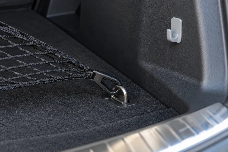 Gros plan sur l’angle d’un coffre de voiture : anneau d’arrimage métallique rabattable dans le plancher, crochet plastique d’un filet de coffre accroché dedans, et à côté un crochet adhésif collé sur la paroi en plastique