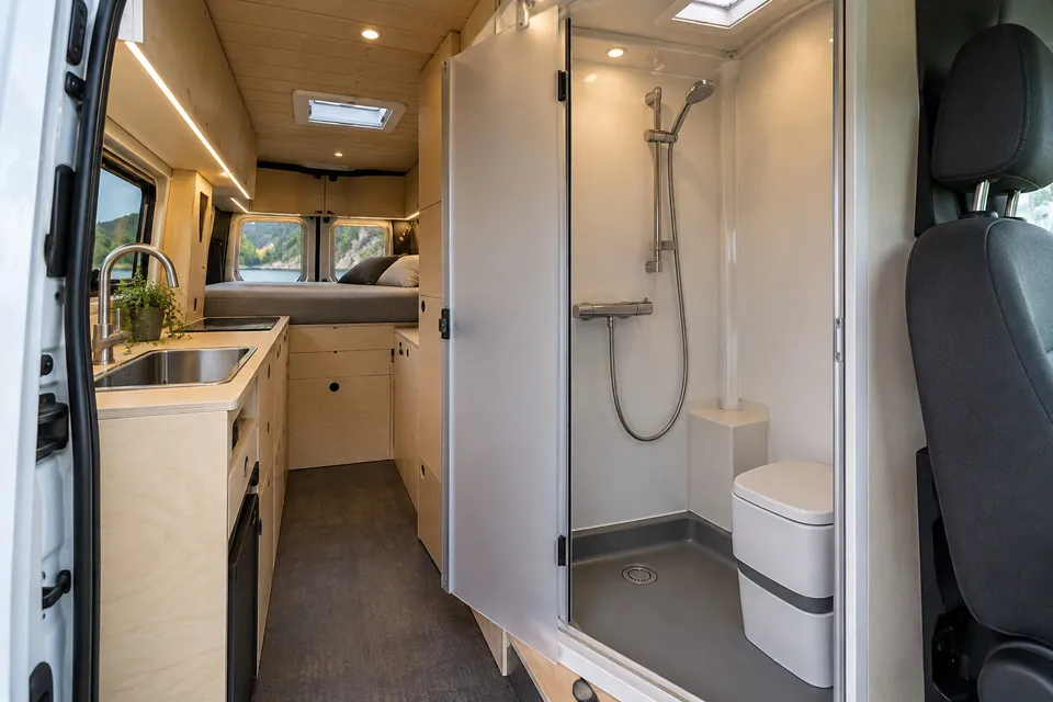 Cabine de douche compacte en kit dans un fourgon aménagé, porte dépolie ouverte sur un bac, un mitigeur et des WC, à côté du bloc cuisine et devant le lit transversal