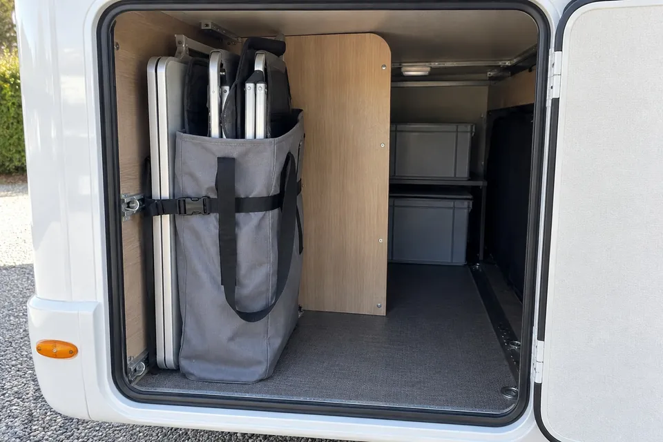 Table de camping pliée et deux chaises pliantes dans une housse commune, debout contre la paroi d’une soute de camping-car près de la porte, maintenues par une sangle et séparées des bacs par une cloison