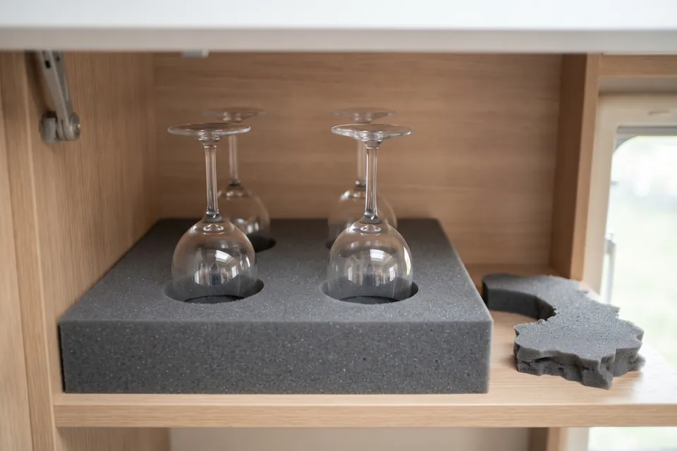 Quatre verres à pied posés tête en bas dans les trous d’un bloc de mousse grise, dans un placard en bois clair de camping-car, avec une chute de mousse découpée à côté